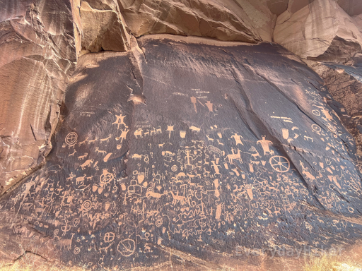 A view of Newspaper Rock in full, displaying a variety of petroglyphs depicting animals; including snakes, sheep, deer, bear paws, turtles/tortoises, antelope, and others, as well as a few figures on horseback and some symbols including at least one apparent wagon wheel.